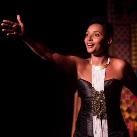 photograph of Mara Menzies. a young black woman with short hair, wearing a stage costume of a black dress and long beaded neck piece. She is performing with her arm outstretched