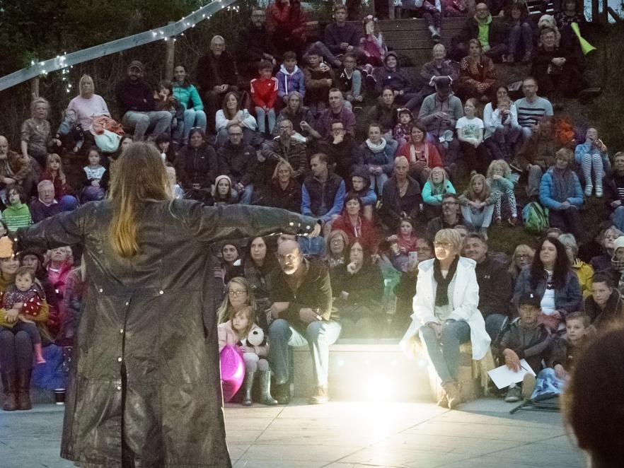 Storytellers are standing on a stage looking out to the audience at night time outdoors.