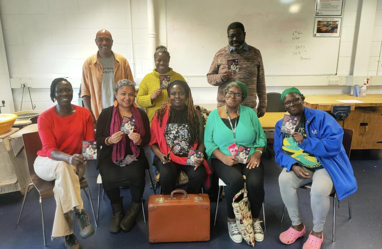 There is a group of people facing the camera and smiling. Some are stood up (back row) and some sat down(front row). They are holding objects that reveal memories and stories.