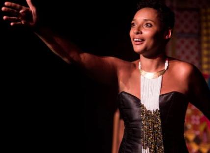 photograph of Mara Menzies. a young black woman with short hair, wearing a stage costume of a black dress and long beaded neck piece. She is performing with her arm outstretched