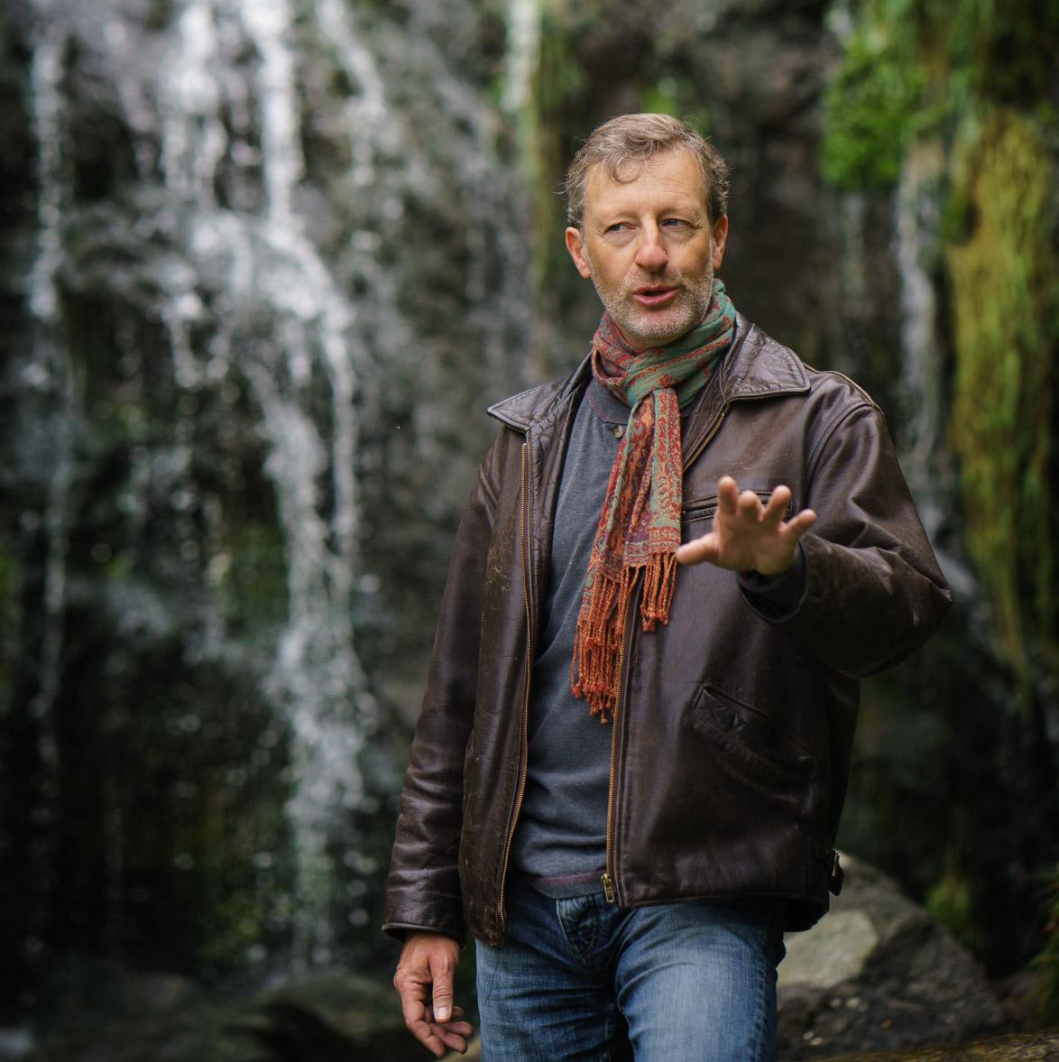 a white man with short brown hair, wearing a brown jacket and red scarf, standing in front of a waterfall