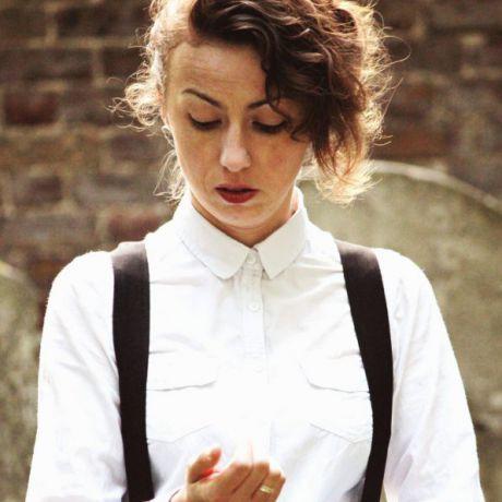 a white woman with long brown hair tied back, wearing a white shirt and black braces