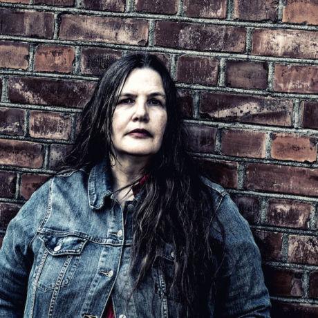 A white woman with long dark hair wearing a denim jacket and leaning against a brick wall