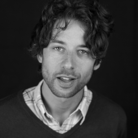 a young white man with curly hair and a beard. wearing a shirt and v neck jumper