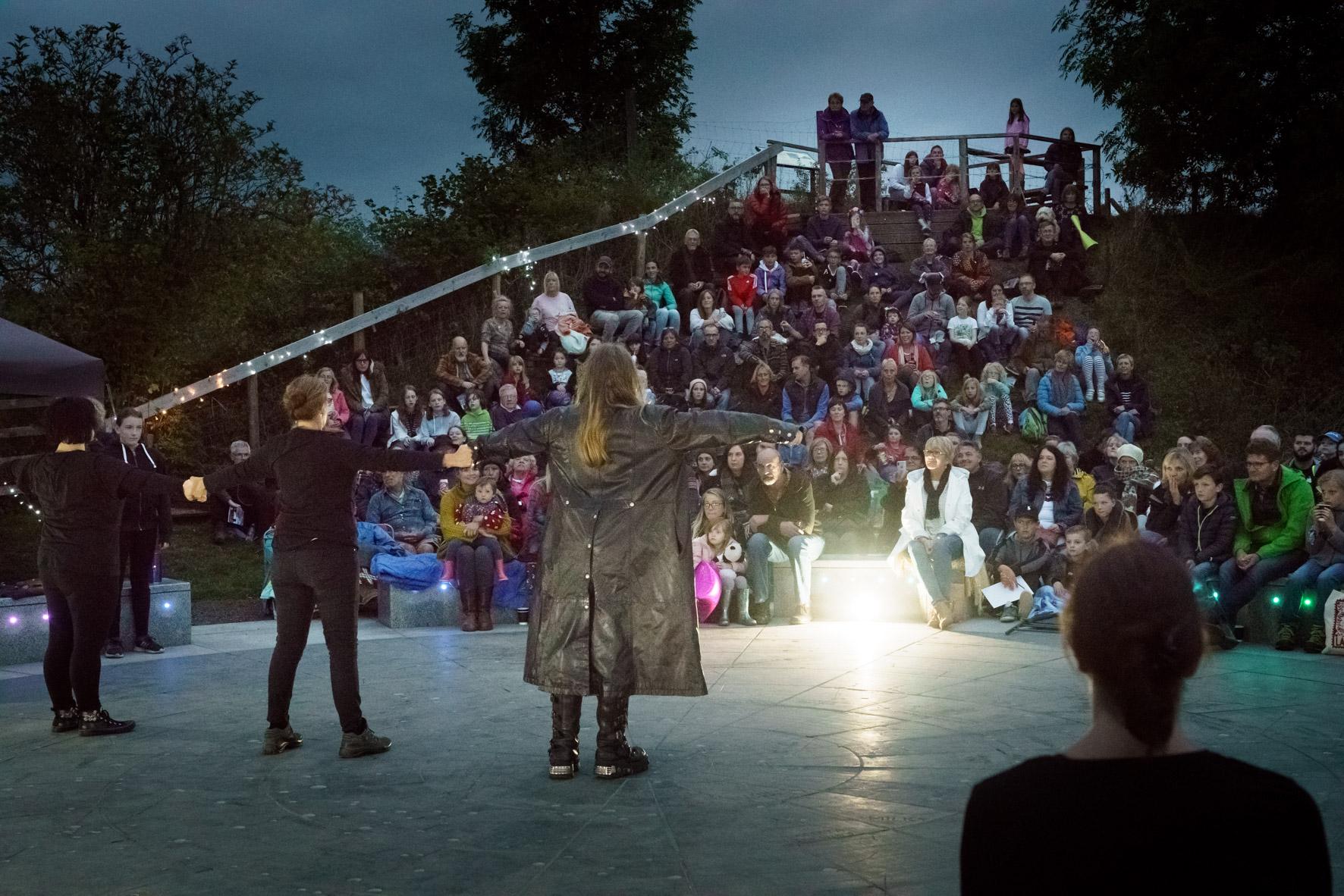 Storytellers are standing on a stage looking out to the audience at night time outdoors.