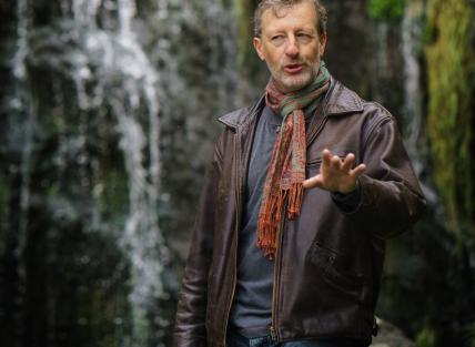 a white man with short brown hair, wearing a brown jacket and red scarf, standing in front of a waterfall