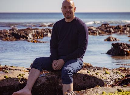 Chris is sat on rocks near the sea.